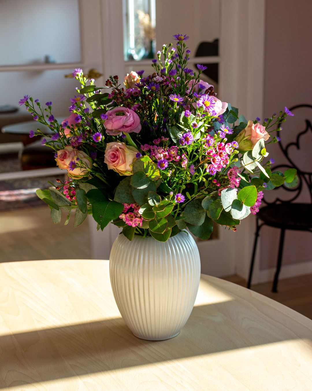 Hvid vase fra Michael Andersen Keramik med blomsterbuket af statice, ranunkler, eukalyptus, roser og astermonarch stående på spisebord i lyst træ