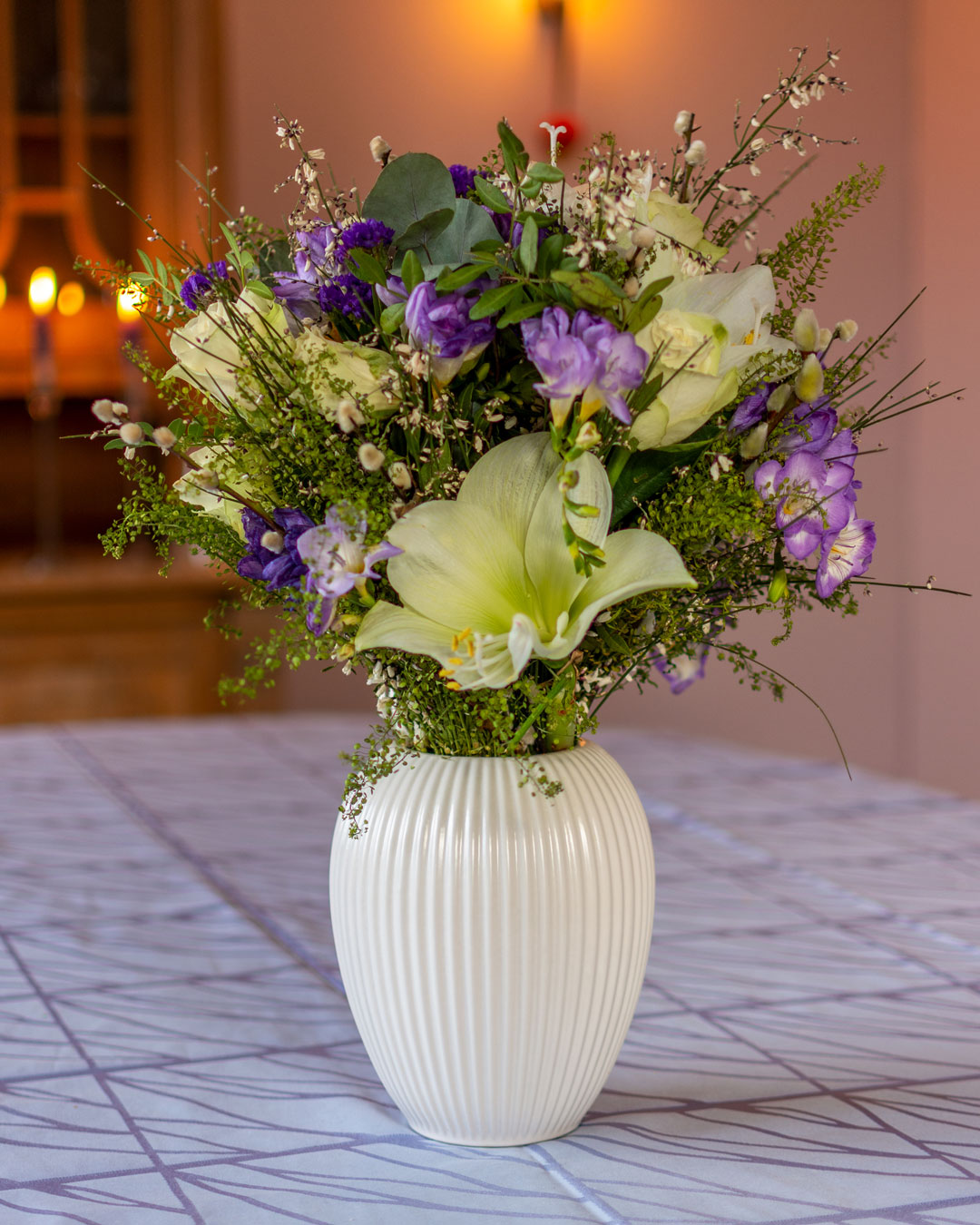 Hvid vase med blomsterbuket af hvide liljer og lilla blomster på lyseblå dug