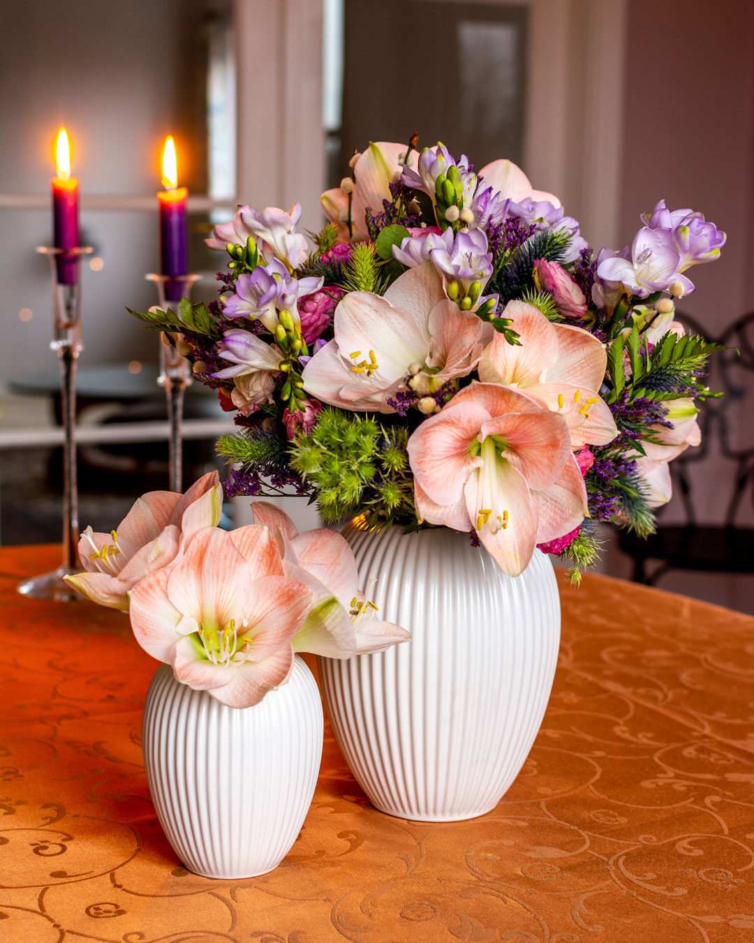 Hvide vaser i lille og mellem størrelse med rosa og lilla blomster på kobberfarvet dug foran lille sterinlys